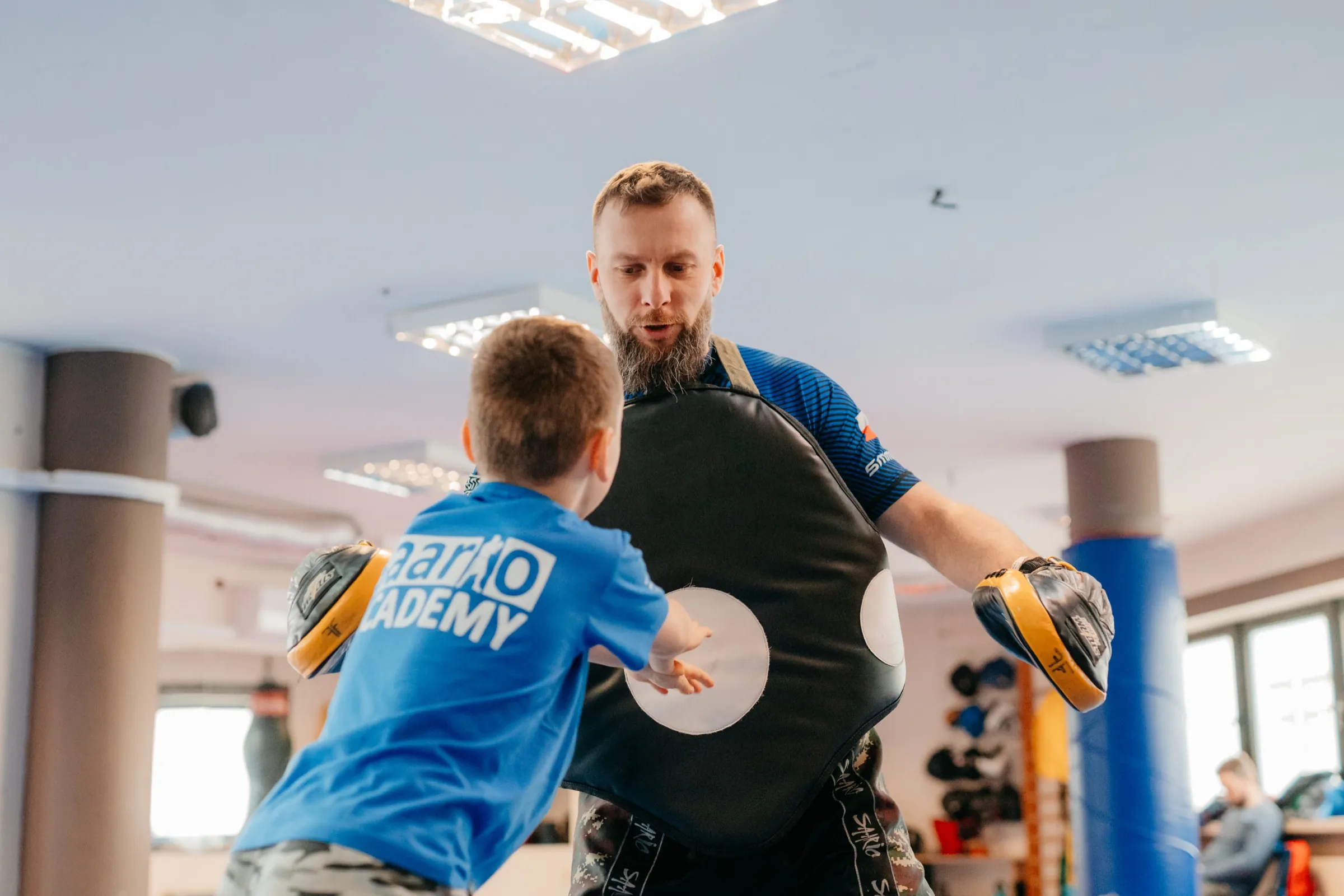 A young boy in a Saario Academy shirt, back of his head to the camera, striking an instructor&rsquo;s body shield on the mats